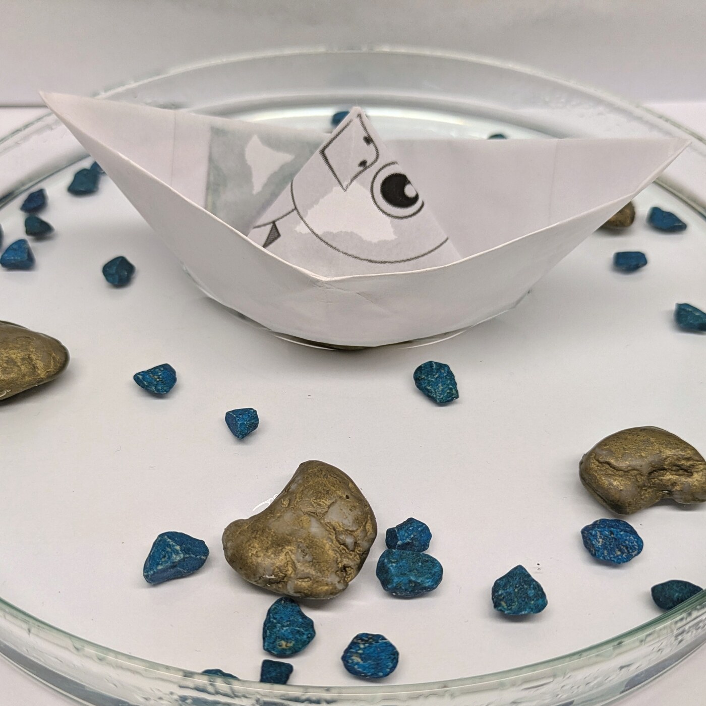 Papierschiffchen in einer Glasschale mit Wasser, Kieseln und blauen Steinen