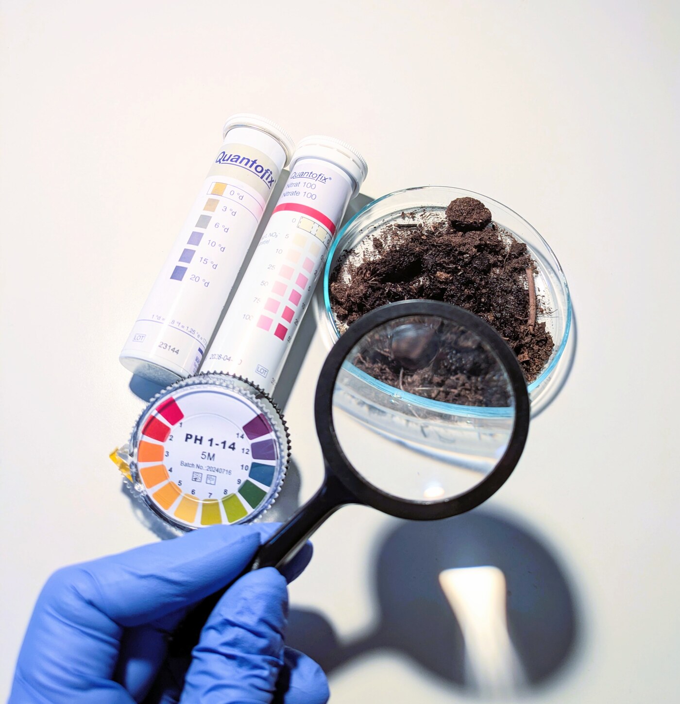 Behandschuhte Hand hält eine Lupe über eine Petrischale mit Erde, daneben Teststreifen für Nitrat und eine pH-Farbskala