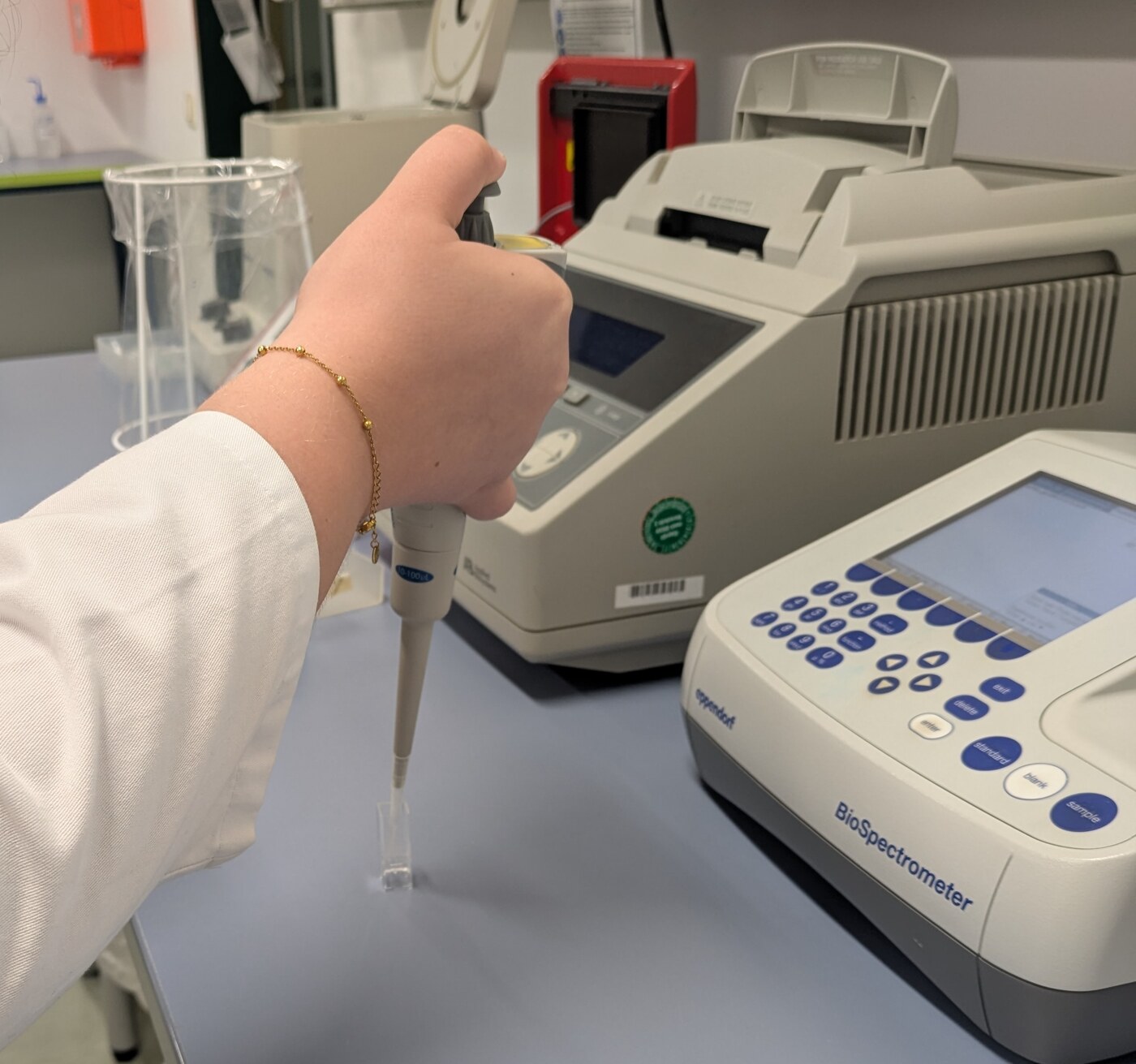 Hand im Laborkittel pipettiert eine Probe, im Hintergrund Thermocycler und BioSpectrometer