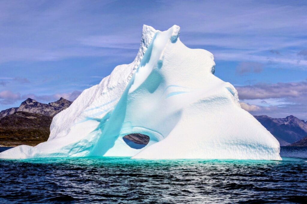 Eisberg mit türkisfarbenen Schmelzrinnen und einer Öffnung im Eis, im Hintergrund Berge